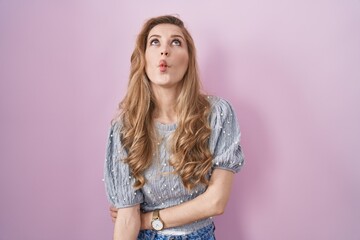 Fototapeta premium Beautiful blonde woman standing over pink background making fish face with lips, crazy and comical gesture. funny expression.