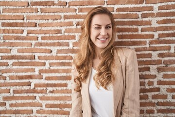 Beautiful blonde woman standing over bricks wall winking looking at the camera with sexy expression, cheerful and happy face.