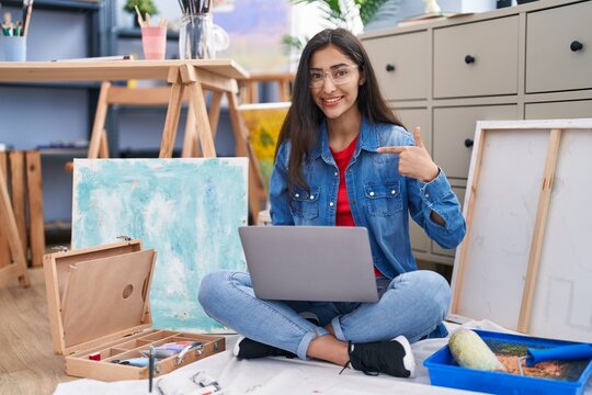 Young teenager girl sitting at art studio using laptop pointing finger to one self smiling happy and proud