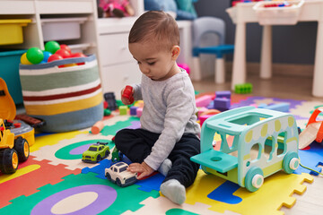 Adorable hispanic boy playing with car toy sitting on floor at kindergarten