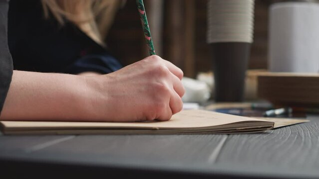 Woman writes in notebook sitting at table in courtyard with wooden buildings. Screenwriter takes notes being at place of historical events on blurred background closeup