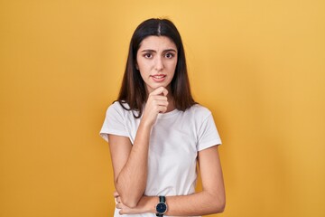 Young beautiful woman standing over yellow background thinking worried about a question, concerned and nervous with hand on chin