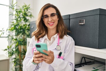 Fototapeta premium Young woman wearing doctor uniform using smartphone at clinic