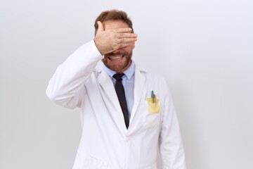 Middle age doctor man with beard wearing white coat smiling and laughing with hand on face covering eyes for surprise. blind concept.