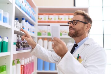 Middle age man pharmacist holding product on shelving reading prescription at pharmacy