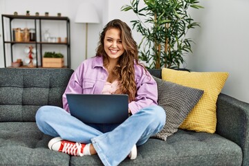 Young beautiful hispanic woman using laptop sitting on sofa at home