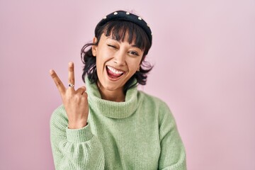 Young beautiful woman standing over pink background smiling with happy face winking at the camera doing victory sign with fingers. number two.