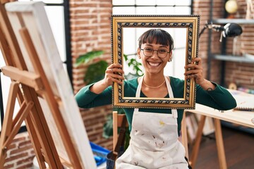 Young beautiful woman sitting at art studio with empty frame smiling with a happy and cool smile on...