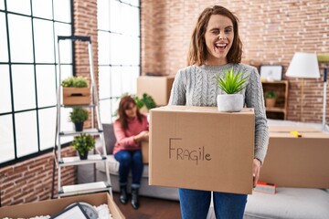 Mother and daughter moving to a new home holding cardboard box winking looking at the camera with...