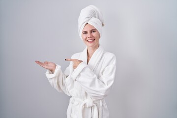 Blonde caucasian woman wearing bathrobe amazed and smiling to the camera while presenting with hand and pointing with finger.