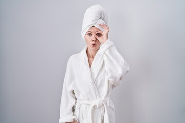 Blonde caucasian woman wearing bathrobe doing ok gesture shocked with surprised face, eye looking through fingers. unbelieving expression.