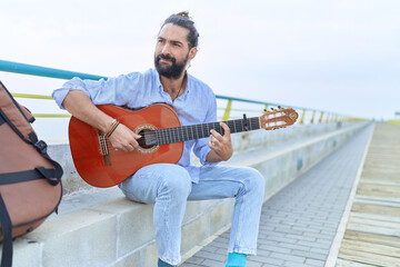 Young hispanic man musician playing classical guitar sitting on bench at seaside