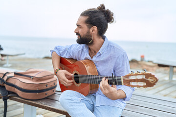 Young hispanic man musician playing classical guitar sitting on bench at seaside