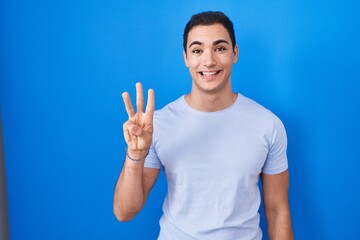 Young hispanic man standing over blue background showing and pointing up with fingers number three while smiling confident and happy.