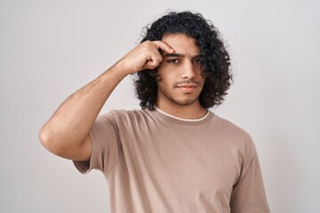 Hispanic man with curly hair standing over white background pointing unhappy to pimple on forehead, ugly infection of blackhead. acne and skin problem