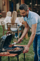 Confident young man barbecuing meat on grill while his family relaxing in the background