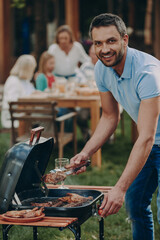 Happy young man barbecuing meat on grill while his family relaxing in the background