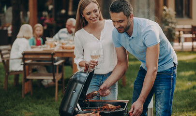 Happy young couple barbecuing meat on grill while their family relaxing in the background