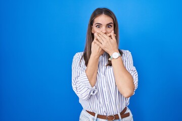 Hispanic young woman standing over blue background shocked covering mouth with hands for mistake....