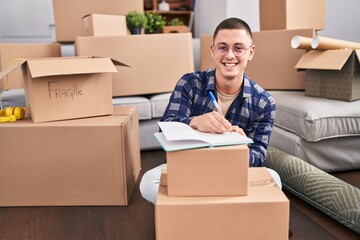 Fototapeta premium Young hispanic man writing on notebook sitting on floor at new home