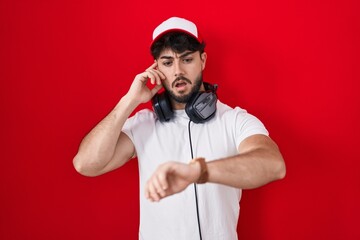Hispanic man with beard wearing gamer hat and headphones looking at the watch time worried, afraid of getting late