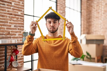 Hispanic man with beard moving to a new home holding ruler puffing cheeks with funny face. mouth inflated with air, catching air. - obrazy, fototapety, plakaty