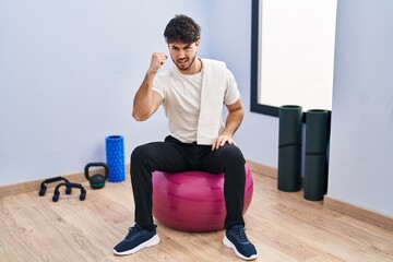 Hispanic man with beard sitting on pilate balls at yoga room angry and mad raising fist frustrated and furious while shouting with anger. rage and aggressive concept.