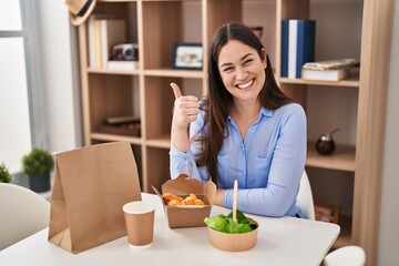 Young brunette woman eating take away food at home smiling happy and positive, thumb up doing excellent and approval sign