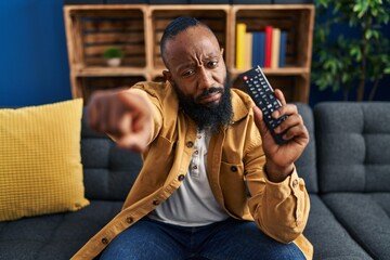 African american man holding television remote control pointing with finger to the camera and to...