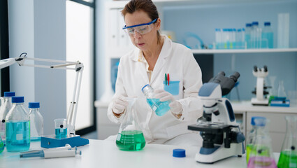Middle age hispanic woman wearing scientist uniform pouring liquid on test tube at laboratory