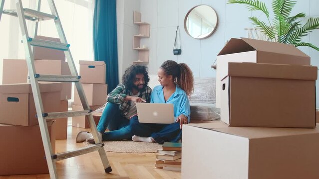 Young happy multiracial couple African American woman and Arabian man with laptop give high five to each other choosing new furniture or interior designer via internet sit on floor near boxes