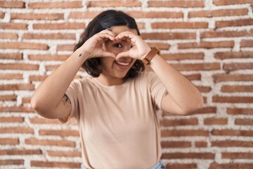 Young hispanic woman standing over bricks wall doing heart shape with hand and fingers smiling looking through sign