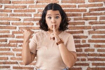 Young hispanic woman standing over bricks wall asking to be quiet with finger on lips pointing with hand to the side. silence and secret concept.