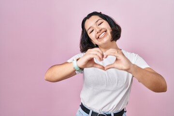 Young hispanic woman wearing casual white t shirt over pink background smiling in love doing heart symbol shape with hands. romantic concept.