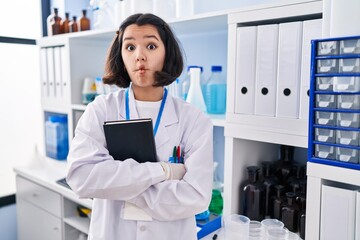 Young hispanic woman working at scientist laboratory making fish face with mouth and squinting eyes, crazy and comical.