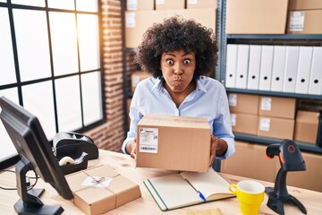 Black woman with curly hair working at small business ecommerce holding box puffing cheeks with...