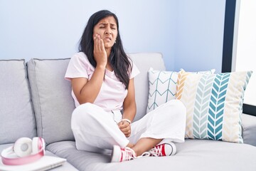 Young hispanic woman sitting on the sofa at home touching mouth with hand with painful expression because of toothache or dental illness on teeth. dentist