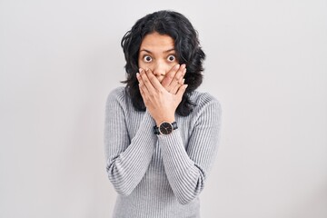 Hispanic woman with dark hair standing over isolated background shocked covering mouth with hands...