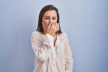 Middle age hispanic woman standing over blue background smelling something stinky and disgusting, intolerable smell, holding breath with fingers on nose. bad smell
