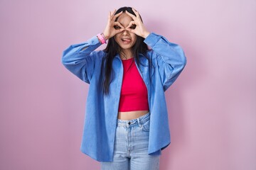 Young asian woman standing over pink background doing ok gesture like binoculars sticking tongue out, eyes looking through fingers. crazy expression.