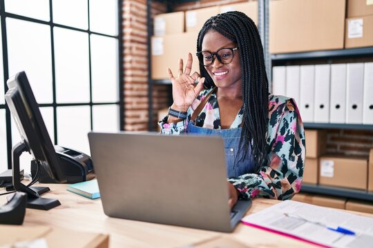African woman with braids working at small business ecommerce with laptop doing ok sign with fingers, smiling friendly gesturing excellent symbol