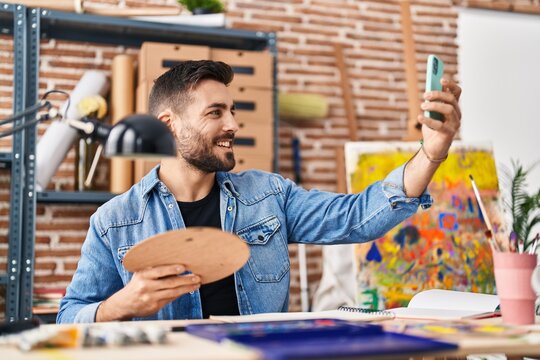 Young hispanic man make selfie by smartphone sitting on table at art studio