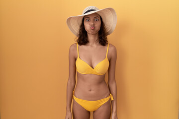 Young hispanic woman wearing bikini and summer hat puffing cheeks with funny face. mouth inflated with air, crazy expression. - obrazy, fototapety, plakaty