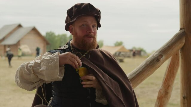Squire with long red beard drinks orange elixir from glass bottle. Hero in medieval clothes replenishes strength using magical liquid closeup on blurred background
