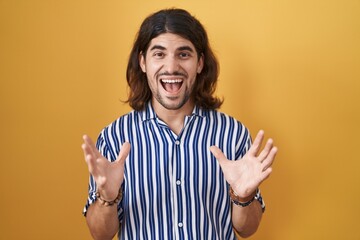 Hispanic man with long hair standing over yellow background crazy and mad shouting and yelling with aggressive expression and arms raised. frustration concept.