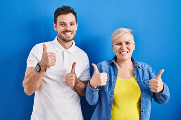 Young brazilian mother and son standing over blue background success sign doing positive gesture with hand, thumbs up smiling and happy. cheerful expression and winner gesture.