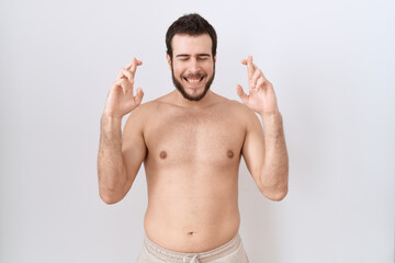 Fototapeta premium Young hispanic man standing shirtless over white background gesturing finger crossed smiling with hope and eyes closed. luck and superstitious concept.