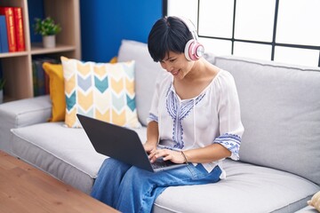 Middle age chinese woman using laptop and headphones sitting on sofa at home