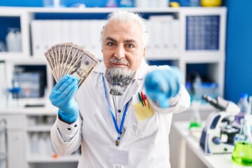 Middle age man with grey hair working at scientist laboratory holding money pointing with finger to the camera and to you, confident gesture looking serious
