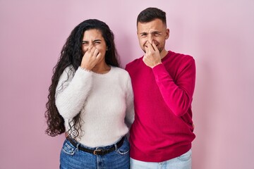 Young hispanic couple standing over pink background smelling something stinky and disgusting, intolerable smell, holding breath with fingers on nose. bad smell